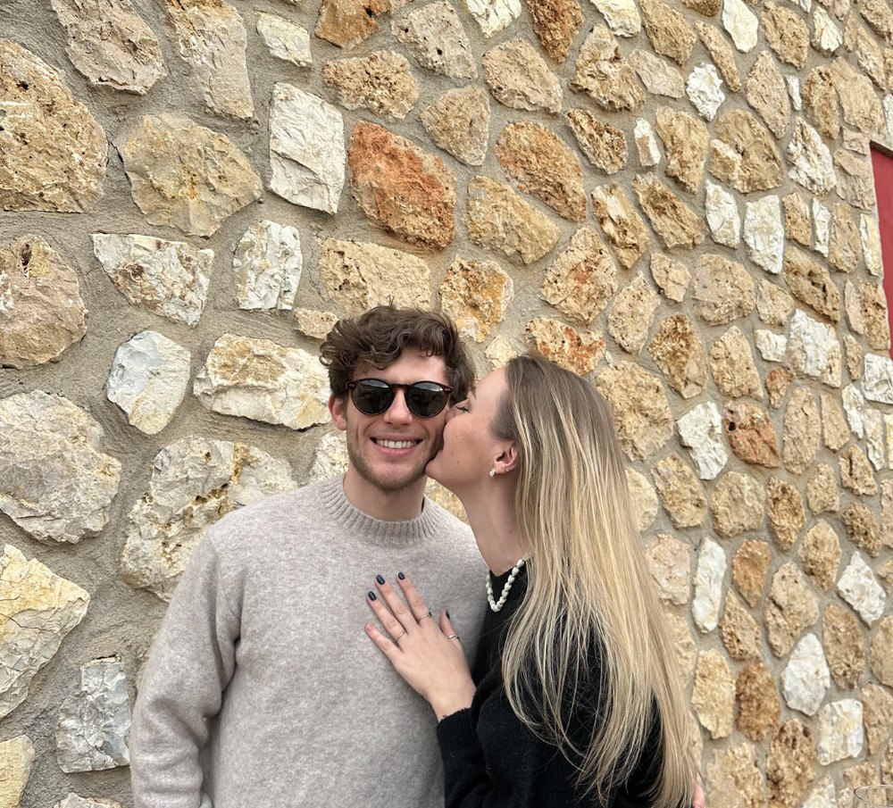 Mona kissing Leonard's cheek in front of a stone wall in Mallorca